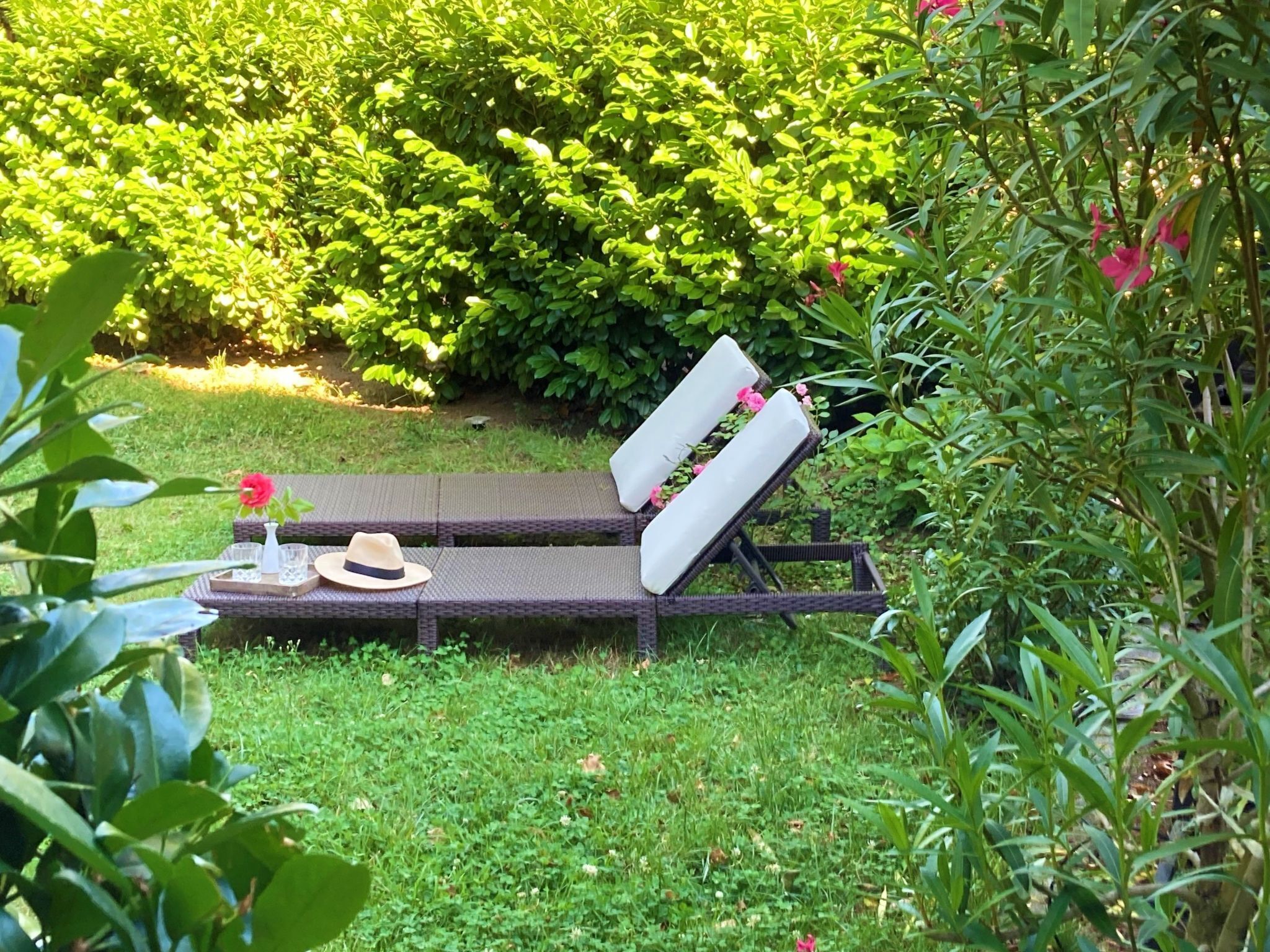 A sun lounger with cushions, a hat, and a tray with glasses and a bottle in a garden setting.