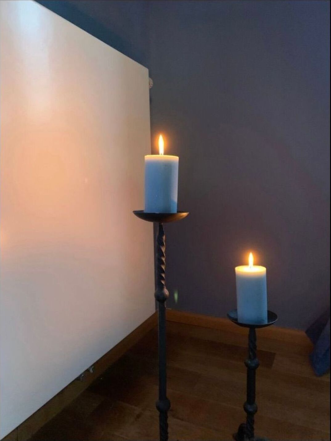 Two lit candles on black candle holders in a room with a plain wall and wooden floor.