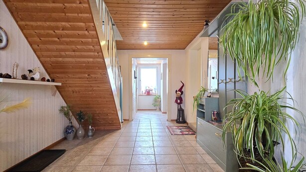 Un pasillo con vigas de madera, suelo de baldosas, una escalera y varias plantas.