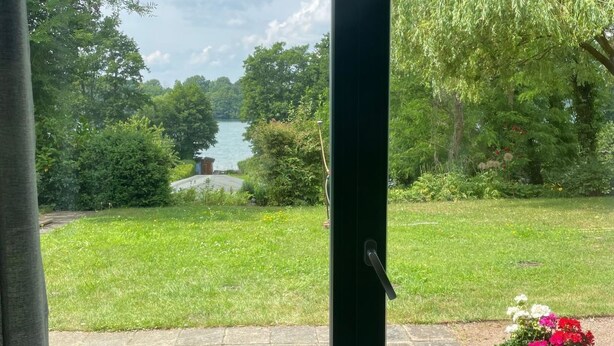 A view through a sliding glass door to a backyard with a wooden deck, a grassy lawn, and a body of water in the distance. - Ferienwohnung Seeweg 4 by Interhome (Lychen)