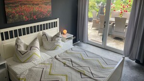 A bedroom with a bed, a nightstand, a wall-mounted lamp, a large window with curtains, and a view of an outdoor patio with chairs and plants. - Ferienwohnung Seeweg 4 by Interhome (Lychen)