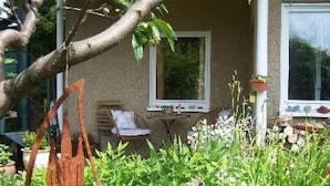 Ein Garten mit verschiedenen Pflanzen und Blumen, ein Stuhl und ein Tisch vor einem Gebäude mit Fenstern.