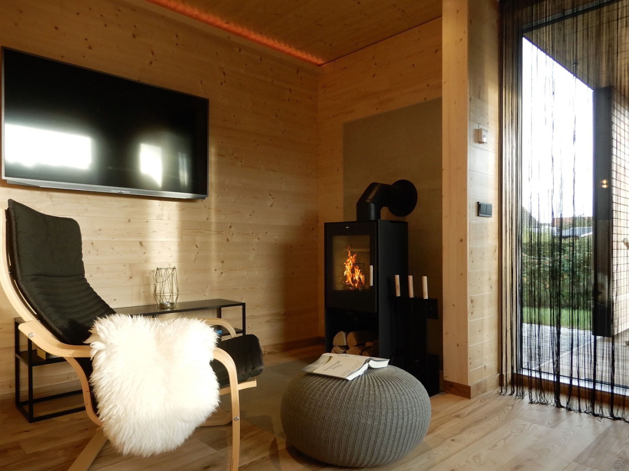 A modern living room with a wooden interior, a black sofa, a white furry ottoman, a glass coffee table, a fireplace, and a large TV mounted on the wall.
