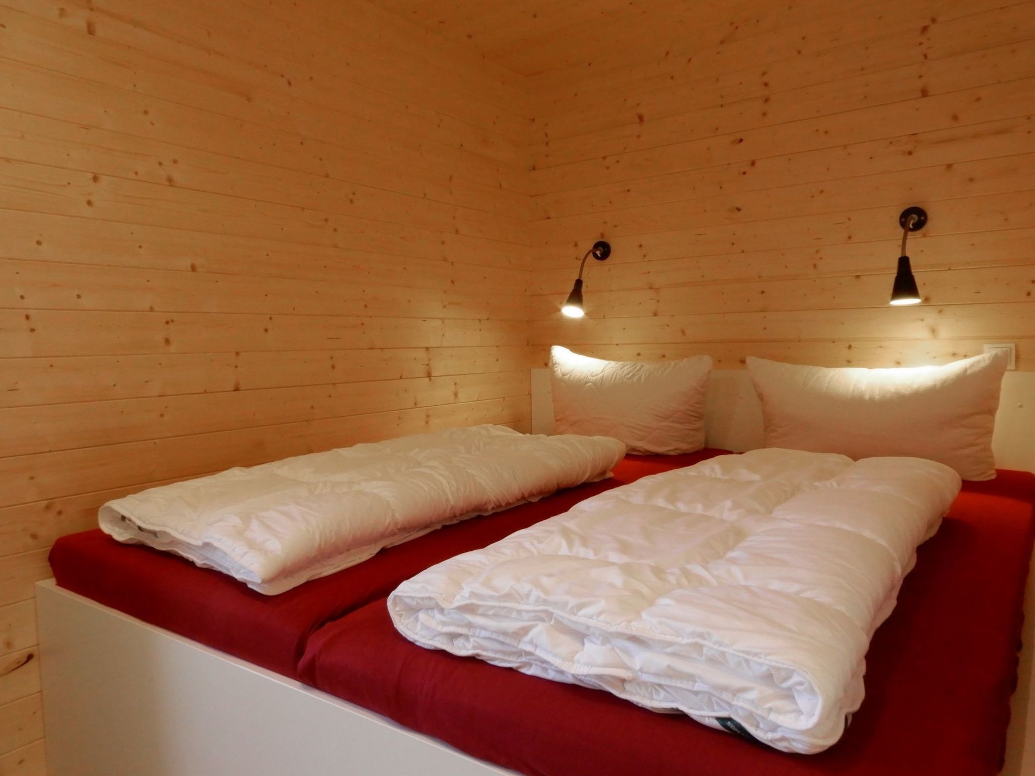 A wooden cabin room with two beds, red bedding, and two wall-mounted lamps.