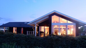 Une maison moderne dotée d’une grande façade vitrée, d’un espace extérieur aménagé et couvert, et d’un toit en pente.