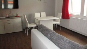 Una habitación moderna y bien iluminada, con una mesa de comedor blanca y sillas, un sofá gris, un televisor de pantalla plana montado en la pared y una ventana con cortinas rojas.
