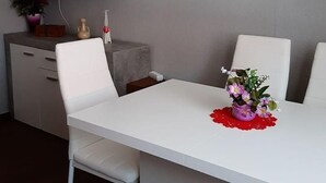 Un comedor moderno con una mesa blanca, dos sillas blancas, un televisor de pantalla plana en la pared, un mueble bajo con un florero y un mantel individual rojo sobre la mesa.