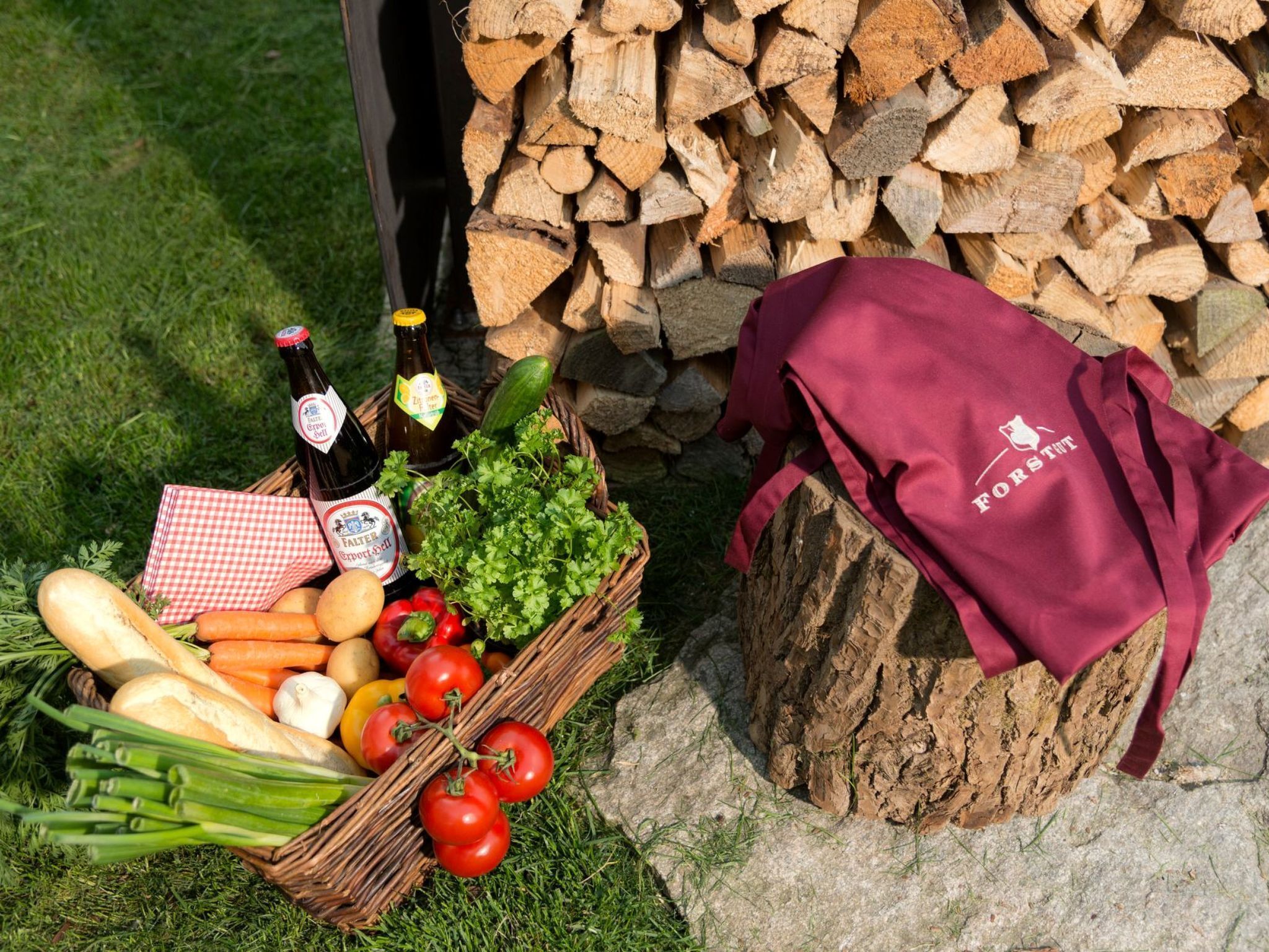 Een picknickopstelling met een mand vol brood, groenten en tomaten, een fles bier en een donkerrode doek met een logo.
