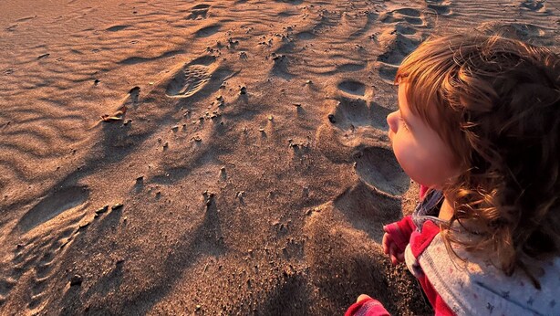 Ein Strandbild mit Fußspuren im Sand und einem Sonnenuntergang im Hintergrund.
