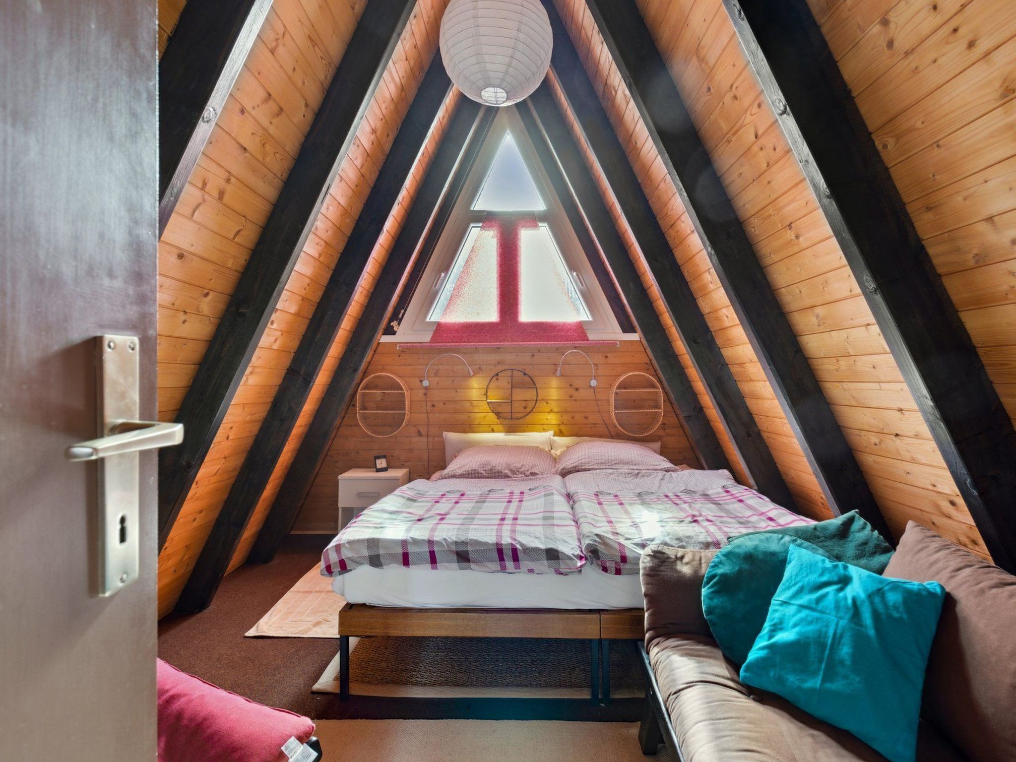 Ein gemütliches Schlafzimmer im Dachgeschoss mit schrägen Wänden, einem Bett mit karierter Bettdecke und einem Fenster mit rotem Rahmen.