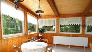 A wooden-paneled room with a round table and chairs, large windows with lace curtains, and a hanging light fixture. - Vacation home Anna by Interhome (Issigau)