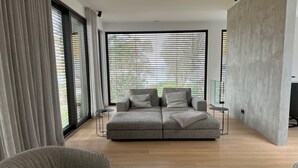A modern living room with a grey sectional sofa, a coffee table with magazines, and large windows with blinds. - Modernes Einfamilienhaus by Interhome (Fahrland)