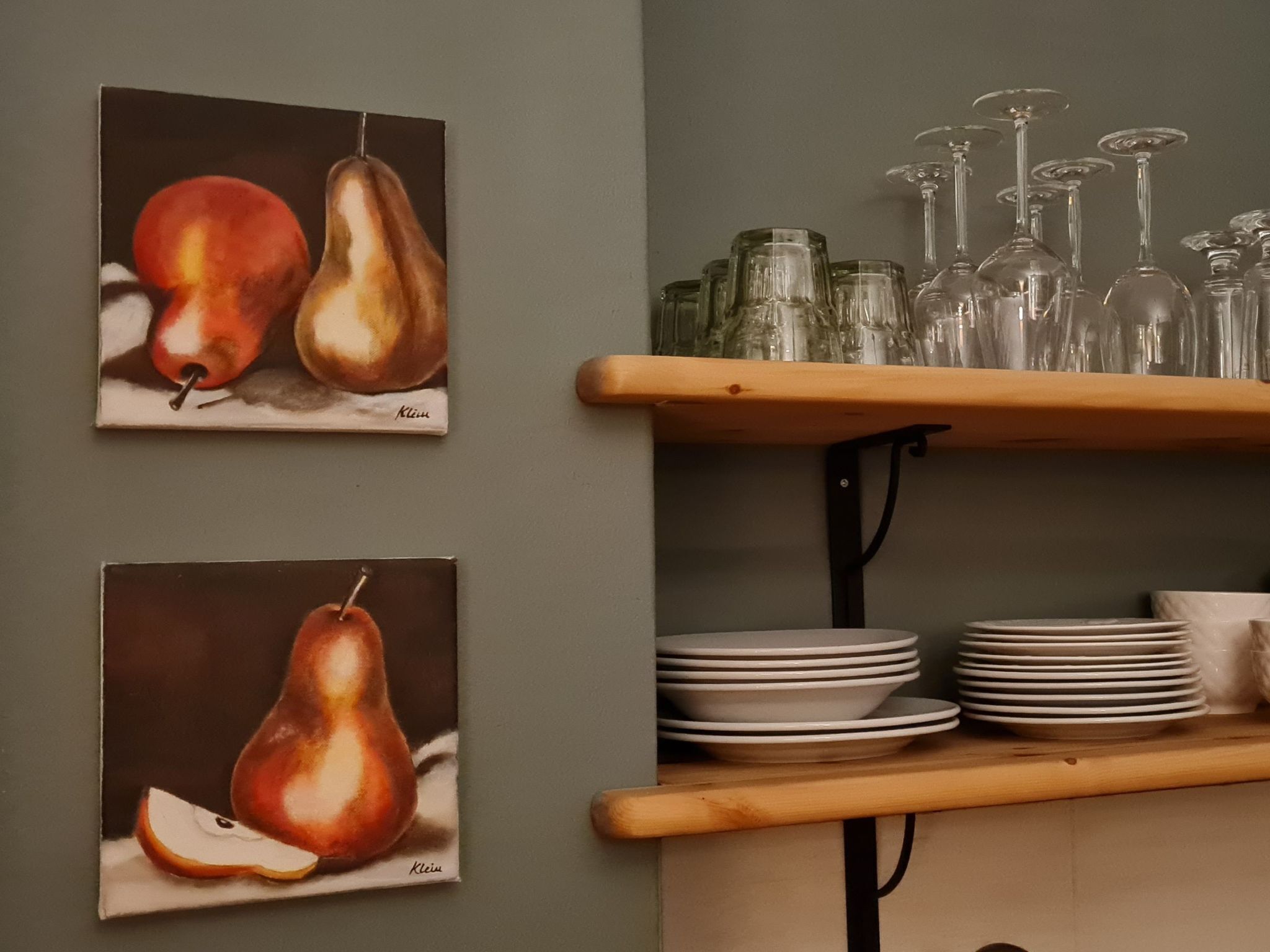 Deux tableaux représentant des poires sont accrochés au mur, une étagère en bois supporte des assiettes et des verres, et une pile d’assiettes est posée sur l’étagère.