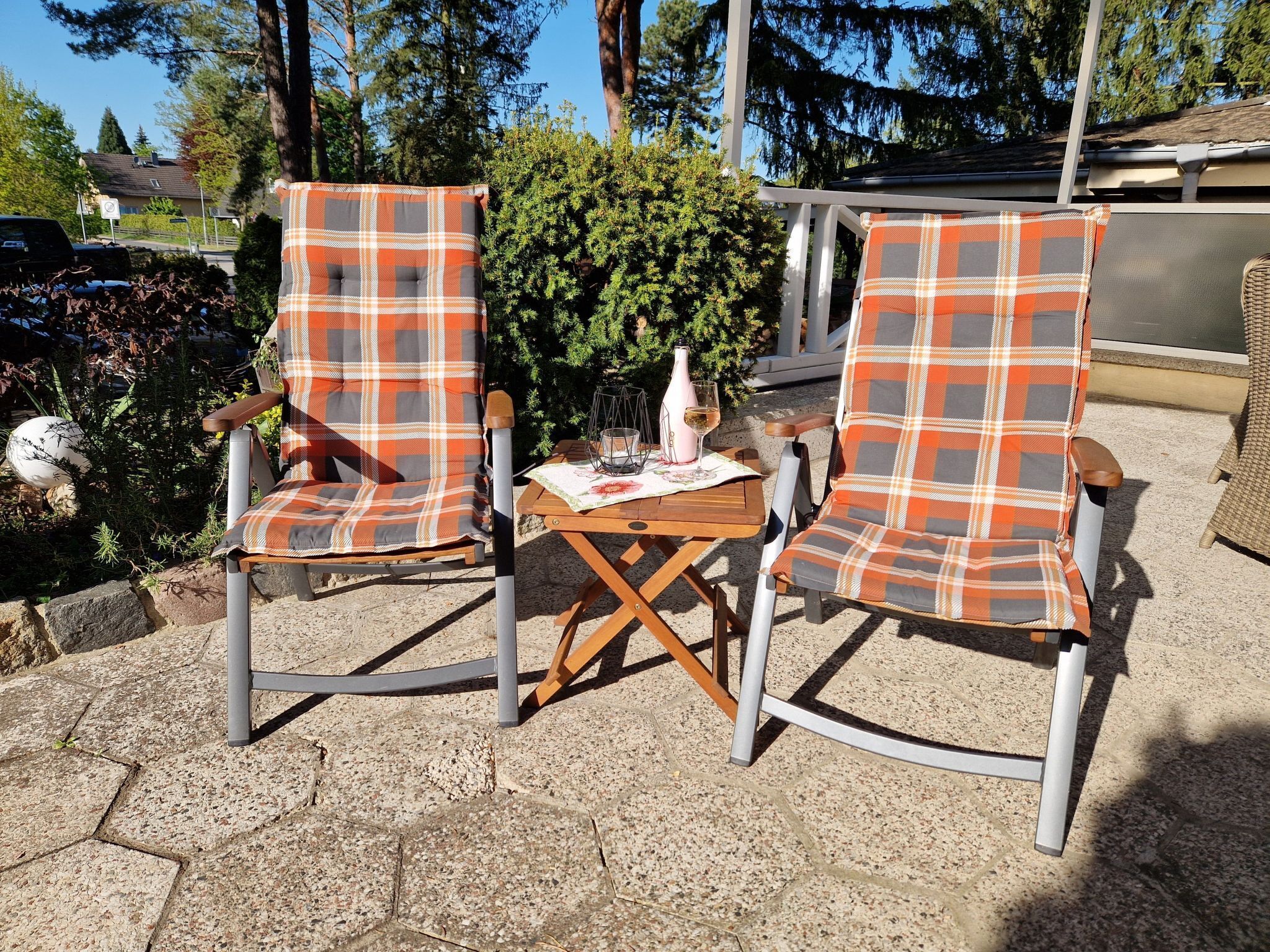 Two plaid chairs with a small table between them, set on a stone patio with a view of trees and a house in the background.