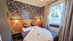 Ein Schlafzimmer mit einem Holzkopfteil, zwei Nachttischlampen, einem Bett mit gesteppter Decke, einem Nachttisch mit gerahmtem Foto und einem Fenster mit Vorhängen.