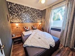 Une chambre à coucher avec une tête de lit en bois, deux lampes de chevet, un lit recouvert d’une couverture matelassée, une table de chevet avec une photo encadrée et une fenêtre avec des rideaux.