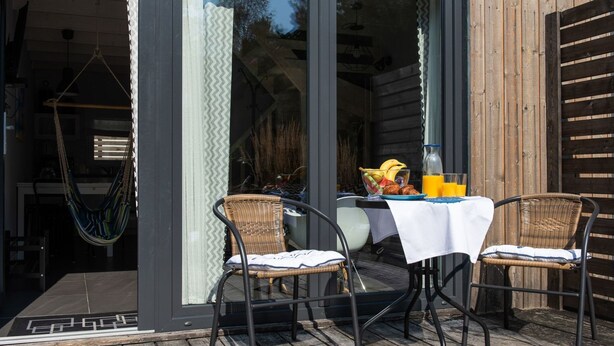 Een terras met twee rieten stoelen en een tafel, gedekt met een fruitschaal, een fles en een glas sinaasappelsap.