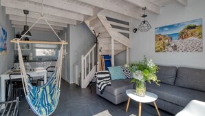 Een moderne woonkamer met een grijze bank, een hangstoel, een eethoek met tafel en stoelen, en een trap naar een ander niveau.
