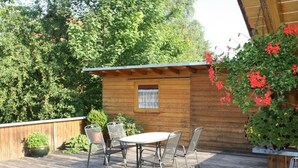 Eine Holzterrasse mit Tisch und Stühlen, umgeben von Grünpflanzen und einer Holzhütte.
