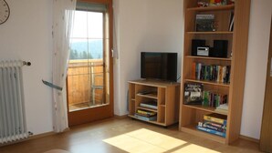 Ein Zimmer mit Holzboden, einem Fernseher auf einem Schrank, einem Bücherregal und einem Fenster mit Vorhängen.