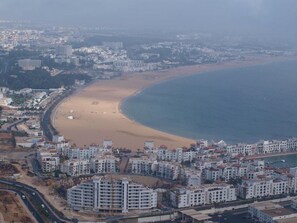 A coastal cityscape with a sandy beach and a large body of water. There are multiple multi-story buildings with balconies, likely residential or hotel properties. - Mogador1 by Interhome (Agadir)