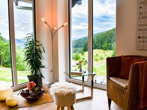 A room with a large window offering a view of a green landscape, a wicker chair, a small table with a glass top, and a white pouf.