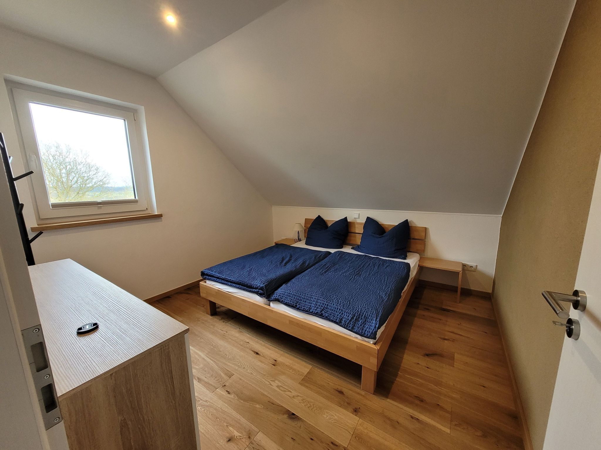 A bedroom with a sloped ceiling, a bed with blue bedding, a wooden desk, and a window with a view of trees.