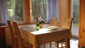 Une table de salle à manger en bois, avec des chaises, une bouteille de vin et une plante en pot, dans une pièce dotée de grandes fenêtres et de murs en bois.