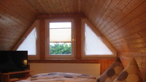 Une chambre confortable située sous les toits, avec un plafond en pente, une grande fenêtre dotée de stores, un lit avec une literie à motifs, une table de chevet avec un téléviseur et une lampe.