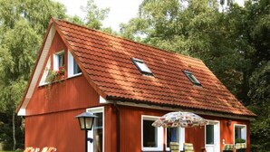 Une maison en bois rouge avec un toit en tuiles, un petit balcon agrémenté de plantes en pot, une terrasse avec des meubles d’extérieur et une piscine.