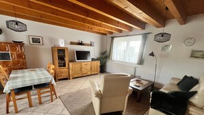 A cozy living room with a wooden ceiling, a dining table with chairs, a sofa, a TV on a wooden cabinet, and a window with curtains. - FH Auf dem Holzweg by Interhome (Bansin)