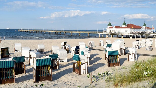 A beach with rows of beach chairs and umbrellas, a pier extending into the sea, and a building with green roofs in the background. - FH Auf dem Holzweg by Interhome (Bansin)