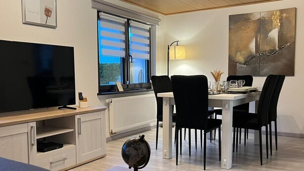 Een moderne woonkamer met een flatscreen televisie, een eethoek met een witte tafel en zwarte stoelen, een wereldbol op een houten standaard, en een schilderij aan de muur.