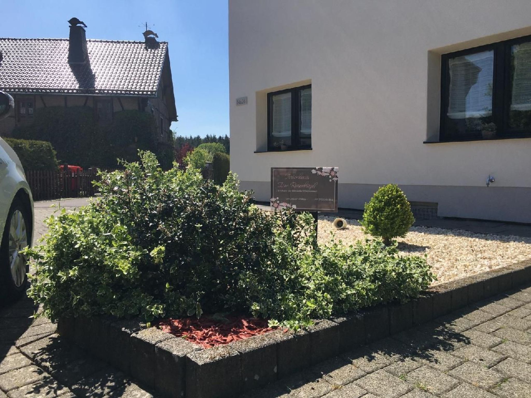 Un edificio con un acceso pavimentado, un pequeño jardín con plantas, una placa conmemorativa y un coche estacionado cerca.