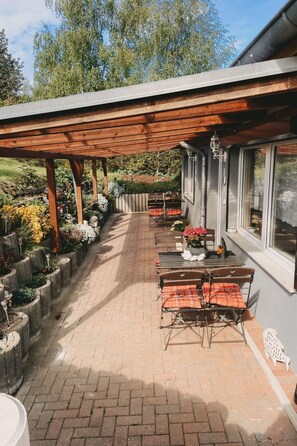 Eine überdachte Terrasse mit Außenbestuhlung, ein Ziegelweg und ein Garten mit verschiedenen Pflanzen.