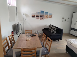 Un salon moderne comprenant un coin repas, un canapé noir et un lit. Trois tableaux représentant des scènes de plein air sont accrochés au mur.