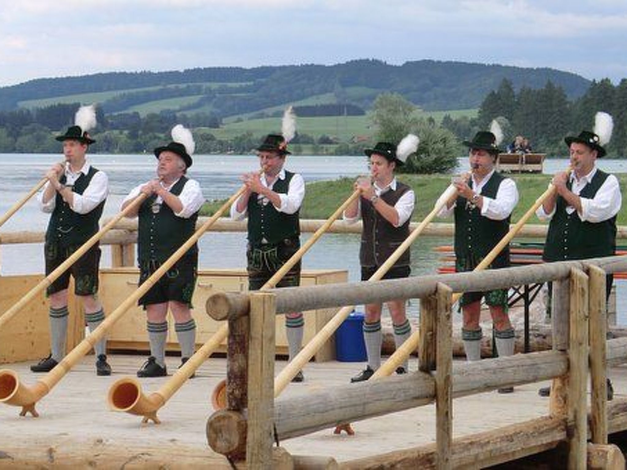 A group of people in traditional attire playing wooden instruments on a wooden platform by a body of water.
