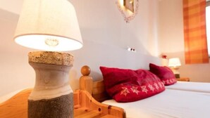 Una habitación con una mesita de noche de madera, una lámpara con pantalla texturizada, una cama con almohadas rojas y una ventana con cortina a cuadros.