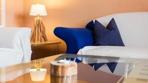 Una habitación de hotel moderna con un sofá blanco, un sillón azul, una pequeña mesita de noche con una lámpara y una mesa de centro de vidrio.