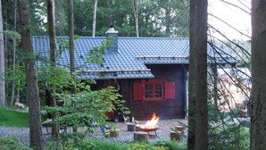 Een rustieke houten hut met pannendak, omringd door bomen en een vuurkuil op de voorgrond.