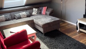 Un dormitorio tipo loft moderno con techo inclinado, un tragaluz grande, un sofá seccional gris, un sillón rojo y una lámpara de pie.