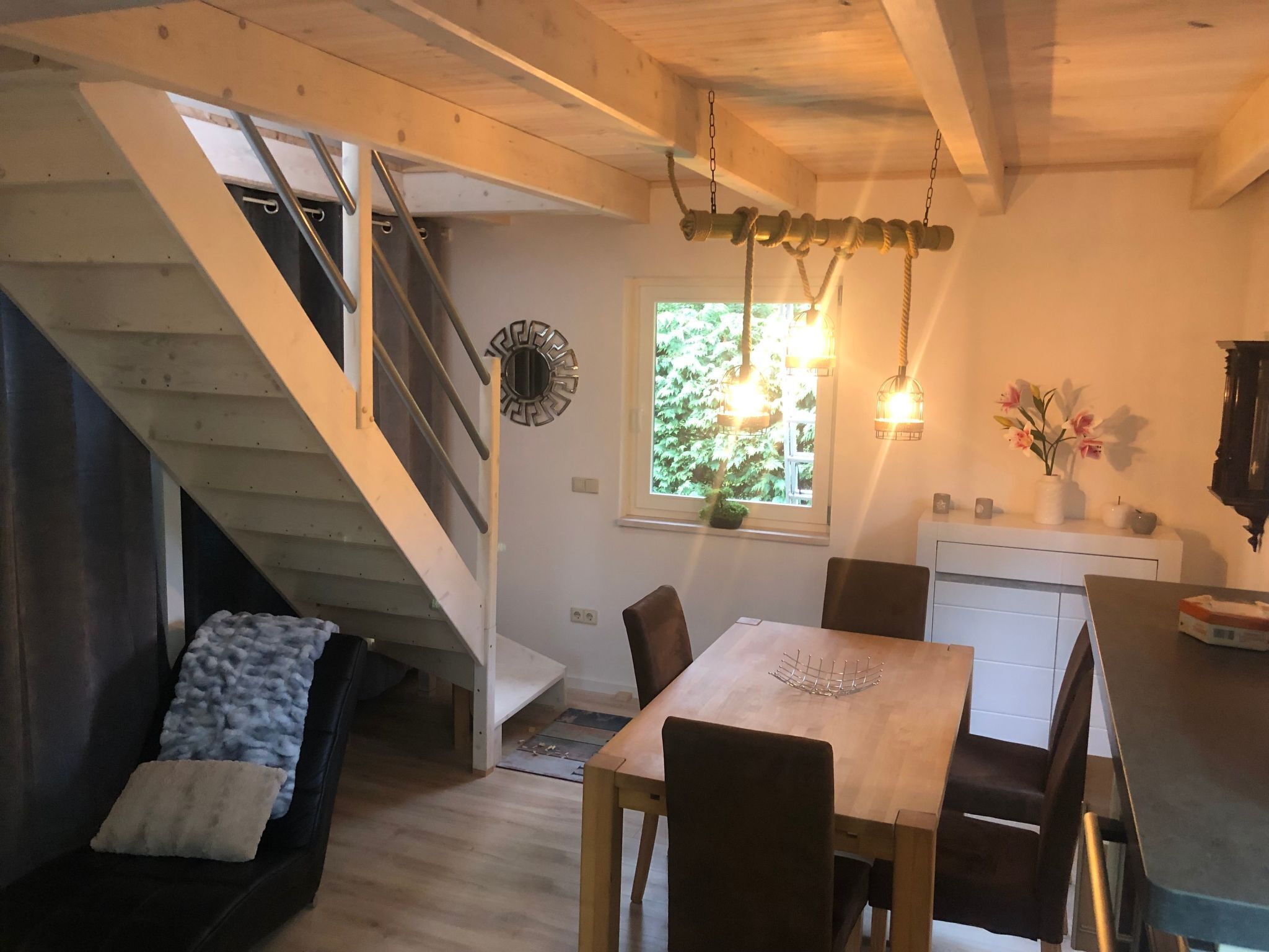 Ein gemütliches Interieur mit einer Holztreppe, einem Essbereich mit Tisch und Stühlen sowie einer Küche mit Schränken und einem Fenster.