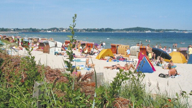 Uma praia com várias tendas e guarda-sóis coloridos, uma faixa de areia e uma vista clara do mar e colinas ao longe.