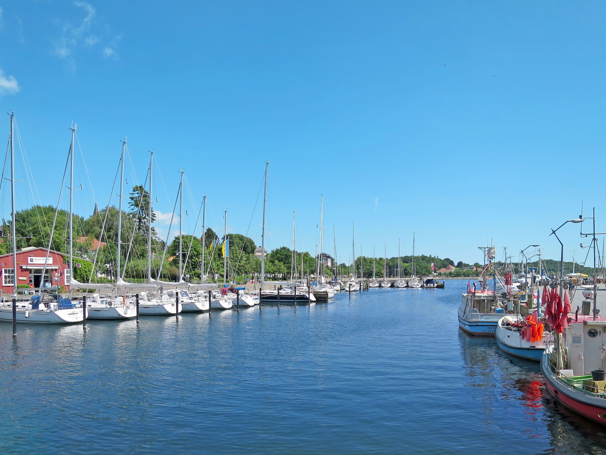 Een jachthaven met meerdere boten afgemeerd, een rood gebouw met de tekst 'Yacht Club' erop, en een helderblauwe lucht.