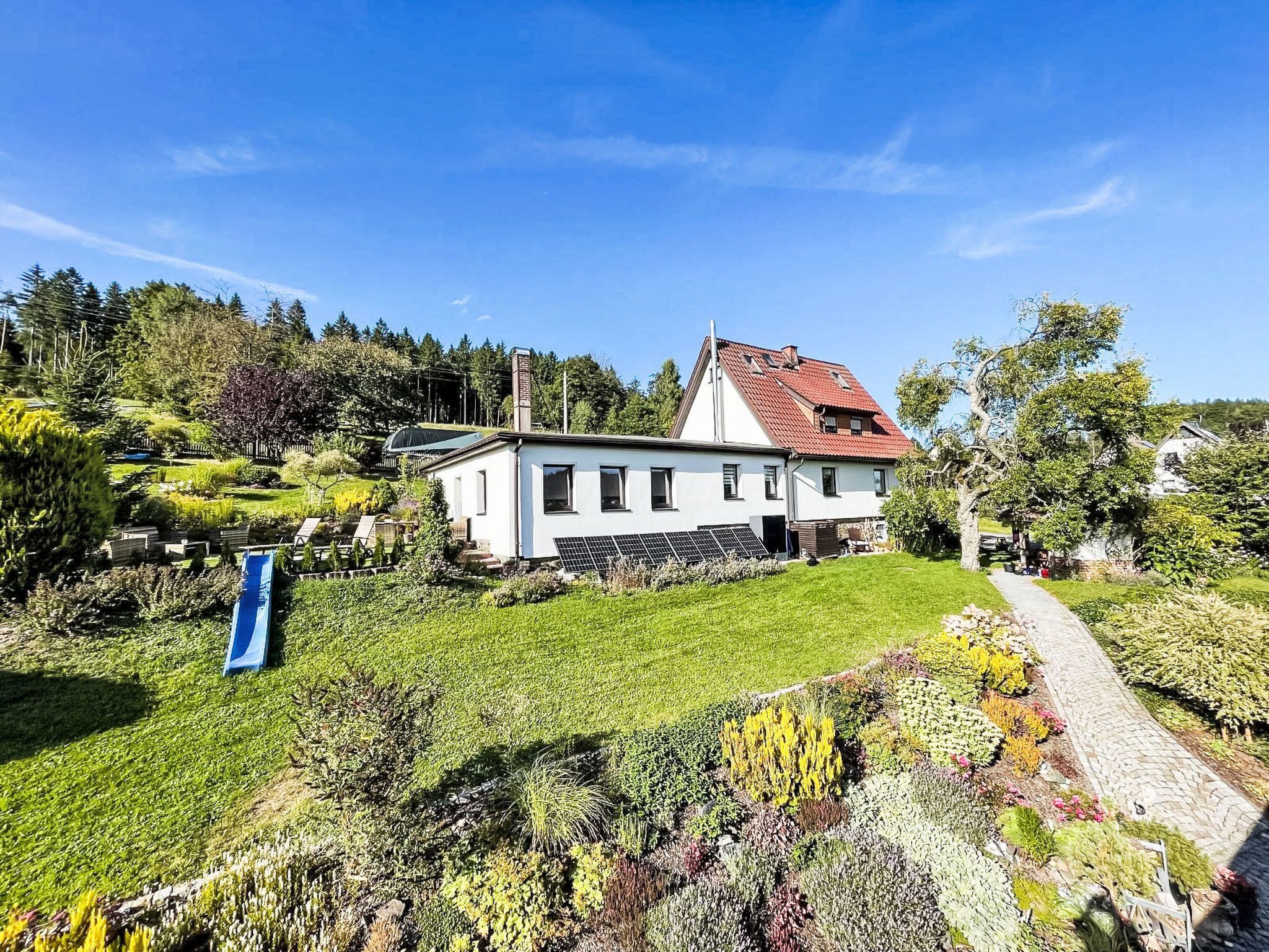 Een wit huis met een rood dak, omgeven door een verzorgde tuin met diverse planten en een blauwe glijbaan.
