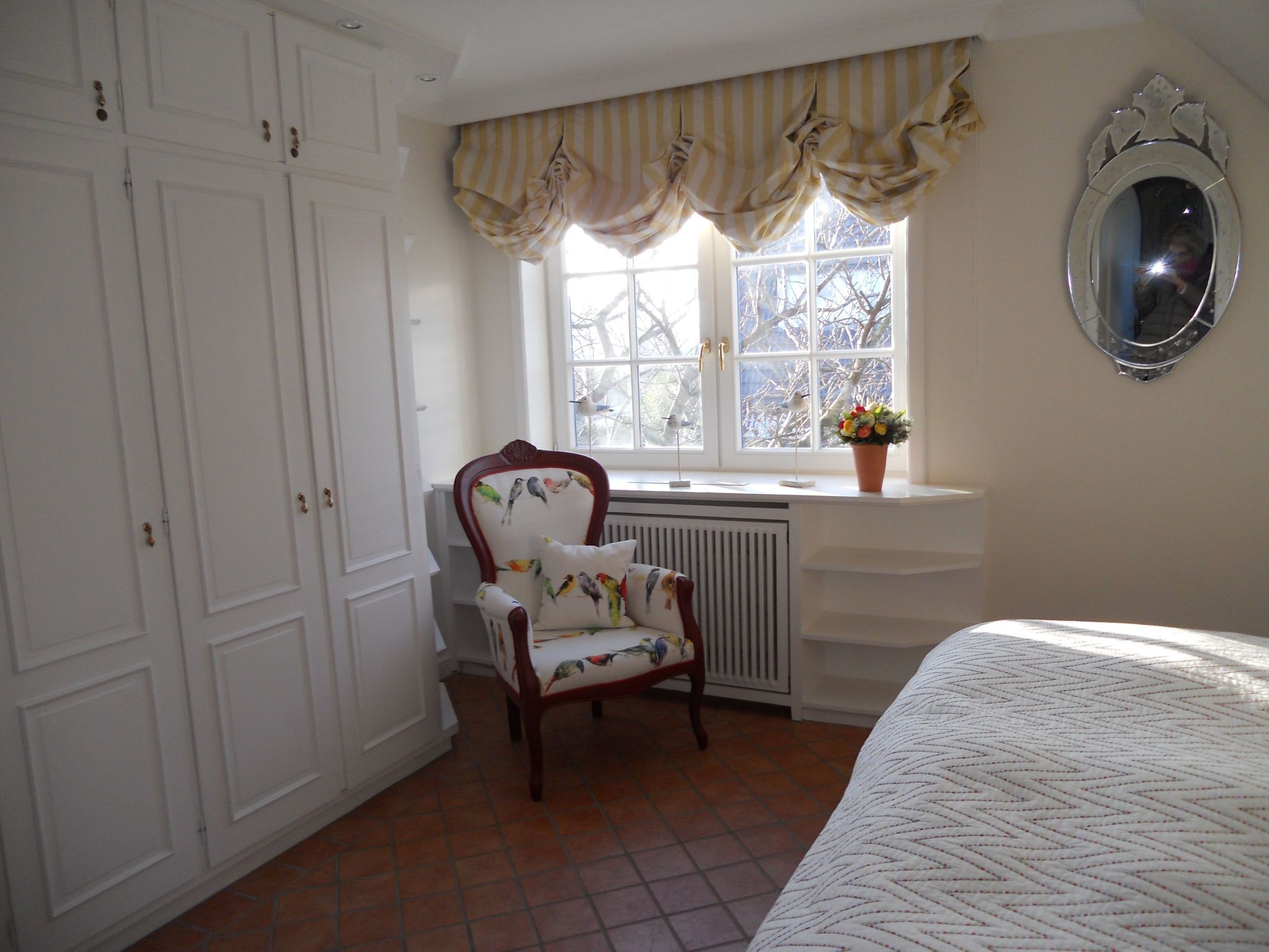 A bedroom with a large window, a radiator, a chair with a floral cushion, a bed with a patterned bedspread, and a mirror on the wall.
