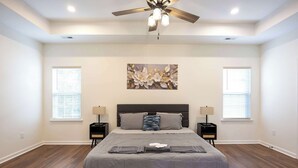 Une chambre à coucher avec un grand lit, deux tables de chevet, un ventilateur de plafond et une œuvre d’art accrochée au mur.