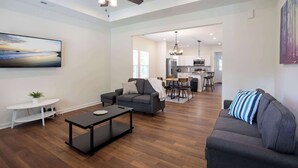 Un salon moderne avec un canapé gris, une table basse et un téléviseur posé sur un meuble blanc. On y trouve également un ventilateur de plafond, un coin repas avec une table et des chaises, et une cuisine à l’arrière-plan.