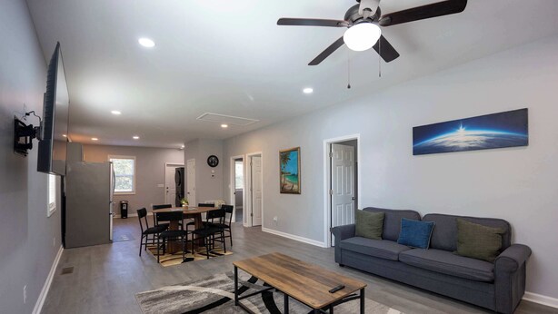 A modern living room with a gray sofa, a wooden coffee table, and a gray rug. There is a dining area with a table and chairs, and a kitchen in the background. - 4 Min to Downtown | 11 to Elon | Car Rental Avail. (Burlington)