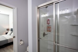 A bathroom with a glass shower door, a toilet, and a bed visible through an open door.